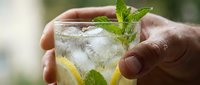 Hands holding a glass of refreshing mojito cocktail with ice, lemon slices, and fresh mint leaves on a blurred green background.