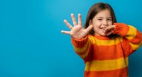 Happy young girl in colorful striped sweater waving hand and smiling on bright blue background. Cheerful child expressing joy and friendliness.