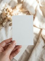 Hand holding blank white card mockup with soft beige fabric and dried flowers background for branding design projects.