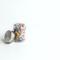 Glass jar filled with colorful rainbow sprinkles and silver lid on white background isolated for baking and dessert design projects.
