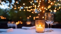 Outdoor evening dinner setup with glowing candle in glass holder and empty wine glass on white table, bokeh string lights in warm golden tones creating ambient atmosphere for celebration or romantic dining event.