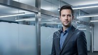 Professional Caucasian businessman in dark suit standing confidently in modern glass office building with sleek architectural elements and corporate interior.
