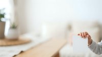 Young child with light skin tone wearing gray long-sleeved clothing holds blank white card in hand against blurred home interior background with wooden table and potted plant.