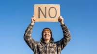 Young adult male with light skin tone wearing plaid shirt and beanie holding cardboard sign with NO text against clear blue sky, expressing disagreement or protest.