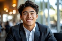 Young Asian professional in black suit and white shirt smiling confidently in modern office setting with soft bokeh background lighting creates welcoming business atmosphere.