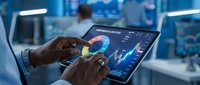 Hands of a male professional with dark skin tone analyzing colorful financial charts and data on a digital tablet in a modern office environment with blue lighting.