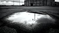 Puddle reflecting cloudy sky on abandoned industrial asphalt ground with deteriorated warehouse building and utility poles in background. Urban decay concept.
