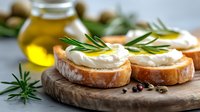 Fresh bread slices with creamy spread and rosemary herbs on wooden board, olive oil bottle in background for gourmet cooking and Mediterranean cuisine.