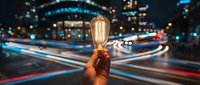 Hand holding vintage Edison bulb against blurred city lights and traffic trails at night, symbolizing innovation and bright ideas in urban environment.