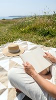 Woman reading book on coastal meadow blanket with straw hat beside her enjoying peaceful outdoor literature moment by ocean with wildflowers and grass.