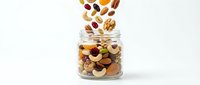 Mixed nuts and dried fruits falling into glass jar on white background, isolated for food packaging, healthy snack marketing, and nutrition content.