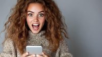 Young Caucasian woman with curly hair wearing knit sweater reacts with excitement and surprise while looking at smartphone screen against gray background.