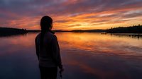 Person standing at lake shore watching dramatic sunset with golden and purple sky reflecting on calm water surface at dusk.