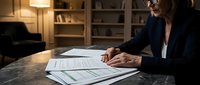 Professional businesswoman reviewing financial documents and reports at modern office desk during evening work session.