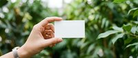 Hand holding blank white business card against lush green tropical foliage background, copy space for branding and identity design.