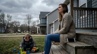 Young Caucasian mother in cozy sweater watching toddler son play with toys on suburban home porch steps during overcast spring day for family lifestyle content.