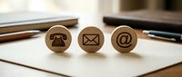 Wooden stamps with communication symbols including telephone, envelope, and email icon on white paper with office supplies nearby.