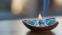 Burning incense in decorative ceramic holder emits gentle blue smoke against blurred background, creating peaceful meditation atmosphere.