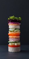 Deconstructed sandwich tower with fresh vegetables, meat and bread layers stacked vertically on dark background for culinary design concepts.