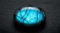 Polished labradorite gemstone with brilliant blue flash and natural mineral patterns on dark textured surface for jewelry and crystal healing applications.