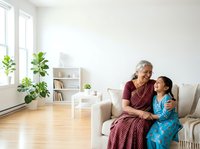 Elderly woman with gray hair and South Asian appearance, wearing burgundy patterned clothing, sitting on beige sofa with young girl in blue dress in bright, minimalist living room with plants and shelving.
