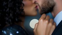 African American woman with curly hair kissing bearded Caucasian man in formal attire against blurred background with bokeh lights, romantic intimate moment captured.