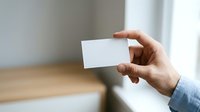 Person with medium skin tone holding blank white business card or paper against blurred interior background, suitable for branding, corporate identity, and marketing materials.
