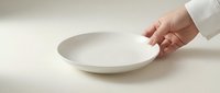 Chef's hand in white uniform holding an empty white ceramic plate on a neutral background, evoking minimalist dining and culinary presentation.
