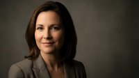 Professional businesswoman with brown hair in gray suit smiling confidently against dark background for corporate headshot and business portrait.