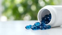 Blue omega-3 fish oil capsules spilling from white bottle on clean surface with green bokeh background for health and wellness supplement concepts.