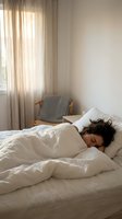Woman with dark hair and medium skin tone sleeping peacefully in bed with white bedding and pillows in a bright, minimalist bedroom with natural light from sheer curtains and wooden chair. Use for wellness, sleep, rest, relaxation, and bedroom design content.