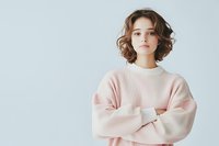 Young woman with short wavy brown hair wearing oversized pink sweater standing with crossed arms against light background, expressing confident casual style.