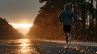 Male runner jogging on wet asphalt road at golden hour sunrise with water splashing from puddles creating dramatic silhouette against warm sunlight filtering through trees.