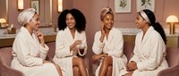 Four diverse women in white bathrobes and towels enjoying spa day together, relaxing on pink sofa in luxury wellness center for self-care and friendship.