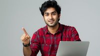 Young South Asian man in red plaid shirt pointing upward while working on laptop against gray background. Professional portrait for business or education content.