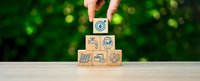 Hand placing wooden cube with recycling symbol on pyramid of eco-friendly icons against green bokeh background for environmental awareness campaigns.