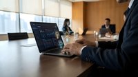Business professional analyzing financial data on laptop during corporate meeting in modern conference room with colleagues discussing strategy.
