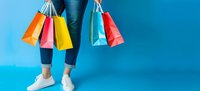 Female legs in jeans and white sneakers holding colorful shopping bags against vibrant blue background. Retail therapy and consumer lifestyle concept.