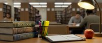 Stack of academic textbooks including biology, world history and literature with laptop, pens and apple on library desk with students studying in background.
