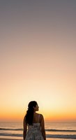 Woman with long dark hair, South Asian ethnicity, wearing a light floral dress, standing alone on beach at sunset gazing toward horizon. Golden and purple sky reflects on calm ocean waves for contemplative lifestyle and travel content.