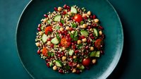 Fresh grain salad with chickpeas, pomegranate seeds, cherry tomatoes, cucumber, and herbs on a teal ceramic plate, top view.