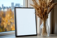 Empty black frame mockup on windowsill with dried pampas grass bouquet in glass vase, autumn city view background, modern minimal home decor.