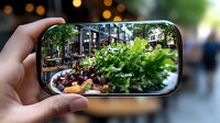 Fresh organic salad with mixed greens and vegetables reflected in smartphone camera lens while taking food photo in outdoor cafe setting with bokeh lights.