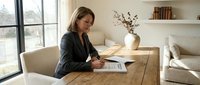 Professional businesswoman working from home office writing in notebook at wooden desk near window with natural light and modern interior design.