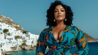 Confident plus size Hispanic woman in turquoise floral dress poses against Mediterranean coastal village backdrop with white buildings and blue sea.