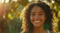 Young African American woman with curly hair smiling outdoors at golden hour, natural sunlight creating bokeh effect through trees, radiating joy and confidence.