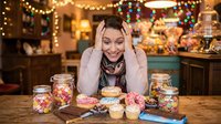 Young woman with long dark hair and fair complexion, wearing a cozy knit sweater, posing cheerfully at a wooden table filled with colorful candies and treats. Warm golden string lights and festive decorations adorn the background, creating a holiday celebration atmosphere perfect for seasonal marketing and lifestyle content.