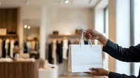 White shopping bag held by person with dark skin tone wearing black sleeve in modern retail clothing store environment for commerce and retail marketing.