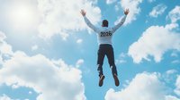 Young Asian man wearing 2024 sweatshirt jumping high against blue sky with white clouds, arms raised in celebration, back view, expressing freedom and success.