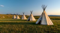 Traditional Native American teepees arranged in circle on grassy prairie landscape at sunset, white canvas dwellings against golden meadow and blue sky background.
